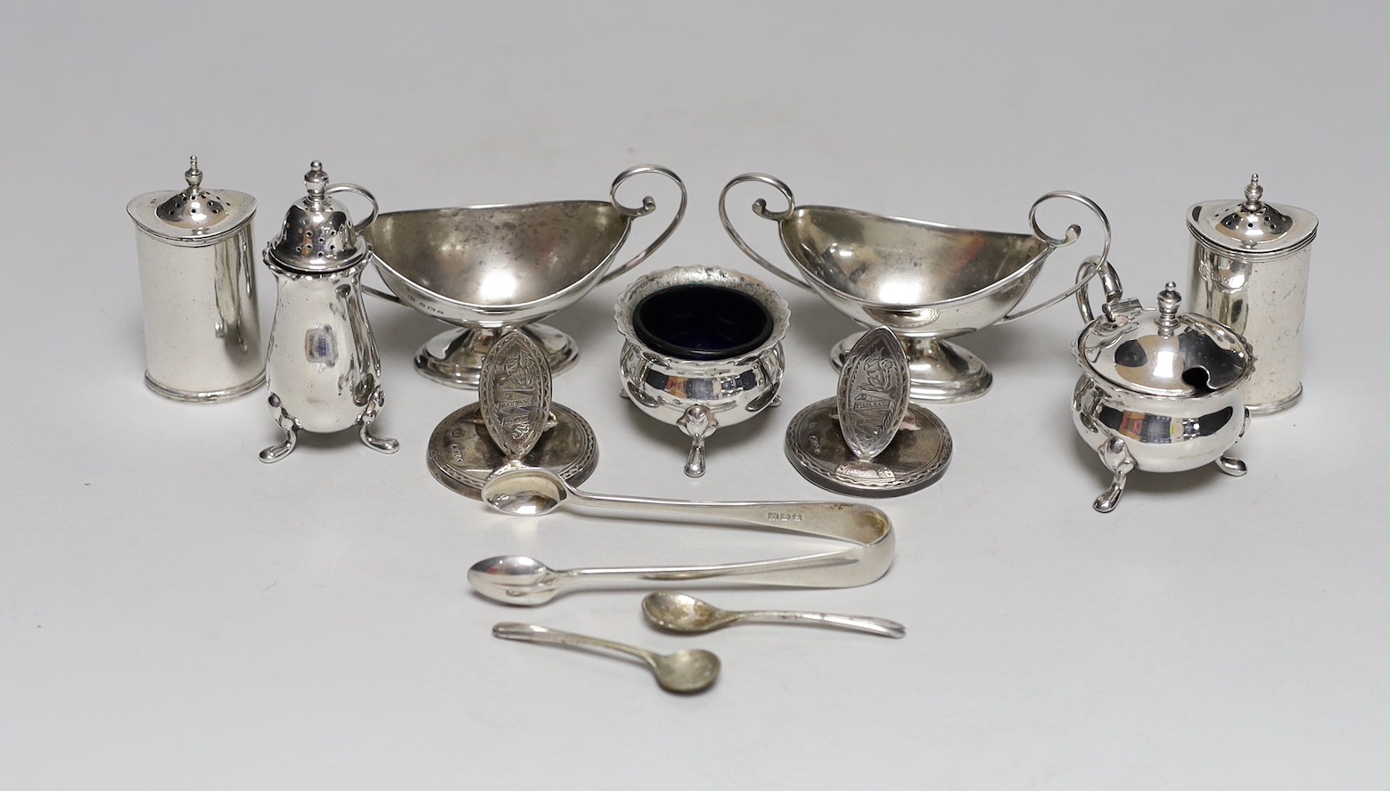 A pair of George V silver two handled boat shaped pedestal salts length 10.7cm, a pair of Edwardian silver pepperettes, a pair of late Victorian silver menu holders, a pair of silver sugar tongs and a three piece plated